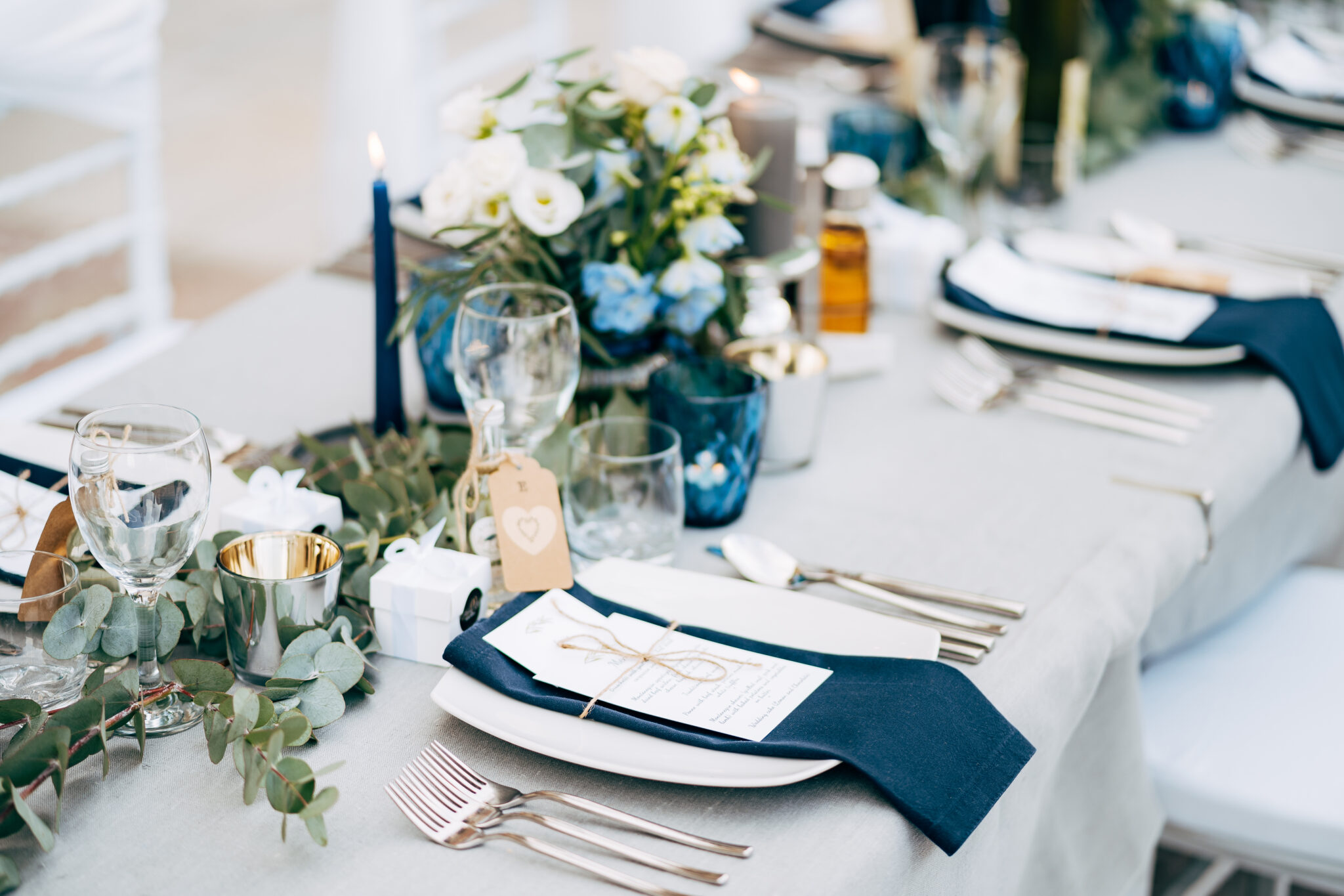 A square plate with a blue cloth towel, knives and forks next to the plate. Flower composition with eucalyptus leaves in the center of the table and burning candles.