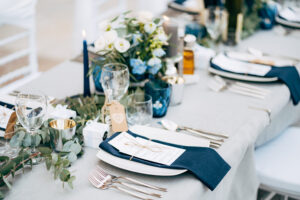 A square plate with a blue cloth towel, knives and forks next to the plate. Flower composition with eucalyptus leaves in the center of the table and burning candles.